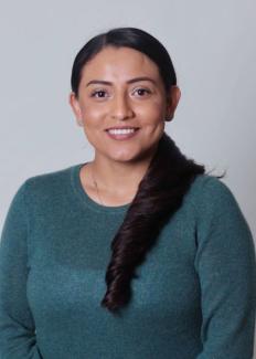 Mariela Aguilar smiling with long dark hair tied in a side braid, wearing a teal sweater against a plain background. The mood is warm and welcoming.