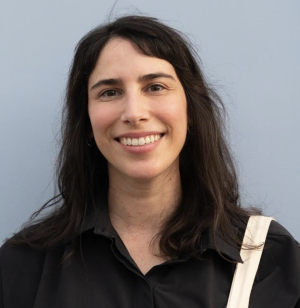 Diana Marcella Vargas with long dark hair smiles warmly, wearing a black shirt and white bag strap against a neutral gray background, conveying a friendly tone.