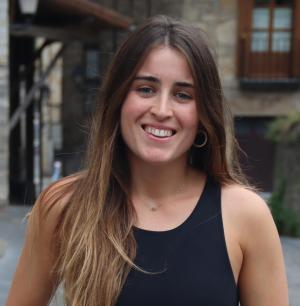 Maitane Muruamendiaraz Arana with long brown hair and hoop earrings smiles warmly, standing outdoors in a cobblestone street setting beside rustic stone buildings.